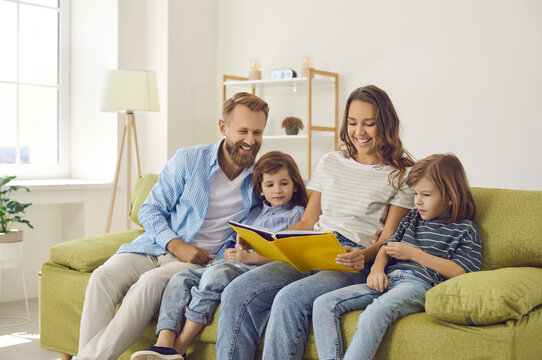 Happy mother, father and children reading book together. Mom, dad and two kids sitting on soft comfy green sofa at home, looking through family photo album or book of interesting stories and fun facts