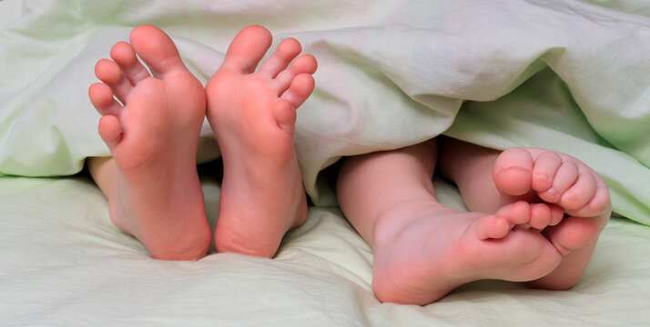 Legs, heels, toes and feet of children lying in bed under a blanket. Close-up of feet of two children, brothers or sisters