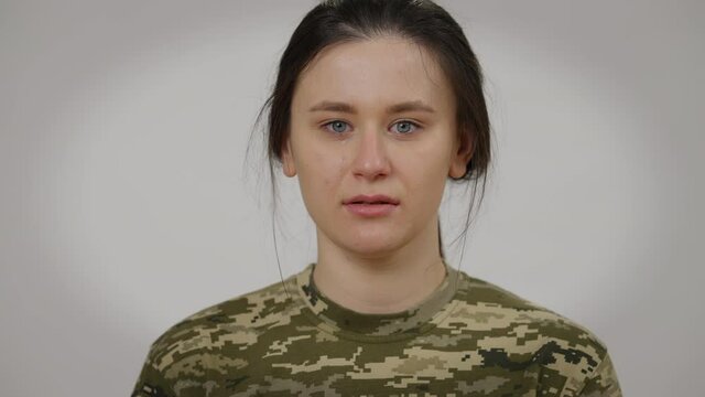 Close-up face of crying female soldier looking at camera. Headshot portrait of sad depressed stressed Caucasian young woman posing at grey background. Overwhelmed recruit cries
