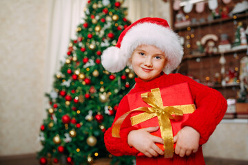 Fototapeta na wymiar A girl in Santa hat holds a gift box with a Christmas gift on the background of a Christmas tree.