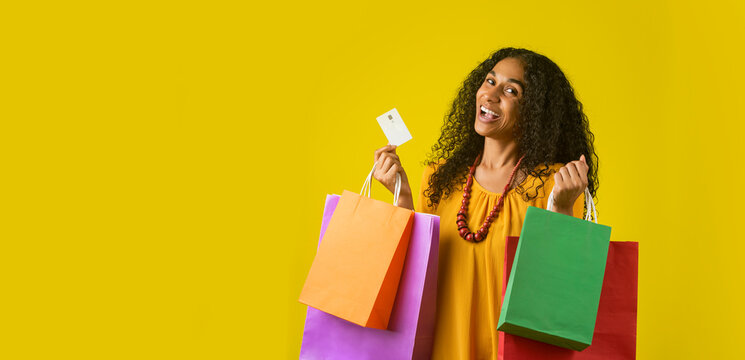 Retrato  de mujer afroamericana alegre en un fondo amarillo sosteniendo bolsas de compra y una tarjeta 