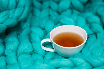 The concept of coziness and comfort is a green knitted blanket on which there is a white cup of tea. Close-up.