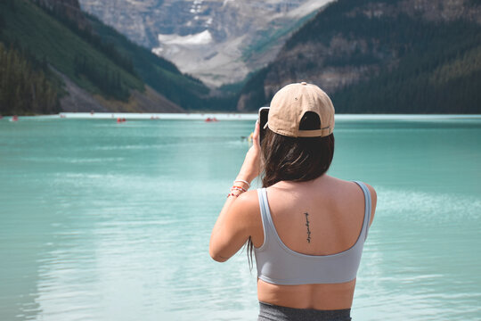 Young girl standing in front of turquoise lake and mountains taking pictures with mobile phone