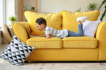 Little African-American boy with laptop watching cartoons while lying on sofa at home