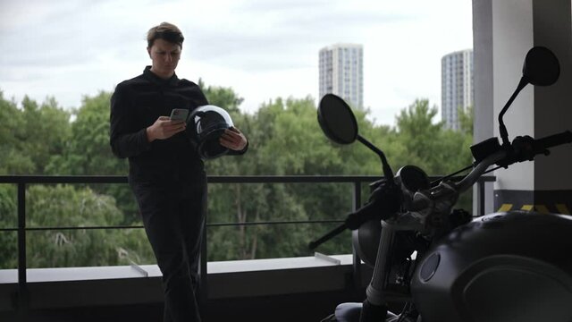 Motorbike driver standing at the parking lot looking at phone, with forest behind him. Young biker looking at phone holding helmet. High quality 4k footage
