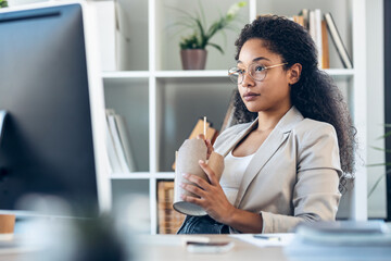 Elegant business woman eating noodles with chopsticks while taking a break of work sitting on chair in the modern startup.