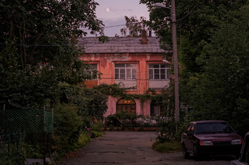 Night outdoor photo of small town old two storey house yard drowned in greenery with an old car parked on the road side in dusk with moon above clouds in dark sky