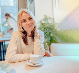 A blonde caucasian joyful girl in a light sweater sits in a restaurant near the window at a table with a cup of cappuccino