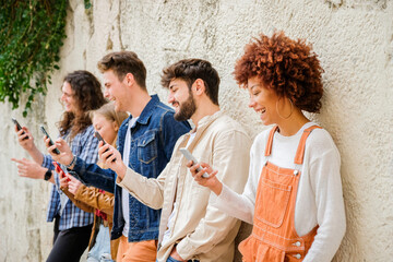 Multicultural group of friends using cellphones outdoors - Students browsing internet holding mobile smart phones standing beside wall