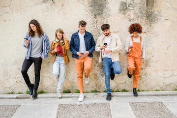 Multicultural group of friends using cellphones outdoors - Students browsing internet holding mobile smart phones standing beside wall