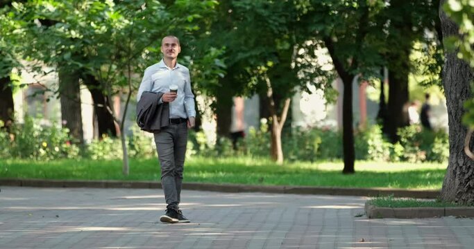 A man with a laptop in his hand and a glass of coffee walks in the park, stops, drinks coffee and looks around. A man walks in a city park.