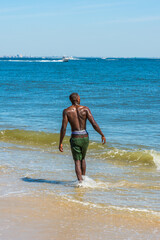 Fototapeta na wymiar A young black guy, half naked, whole body wet, is walking on water of ocean, looking forward. The horizontal is a big city outline, a small boat in the background..