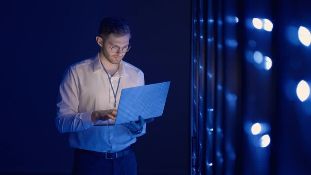 Data center IT specialists coworking in secure server room of cyberspace. Male administrator pointing at error server cabinet running diagnostics on laptop computer