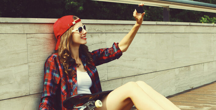 Teenager girl sitting with skateboard taking a selfie by smartphone wearing a baseball cap in the city