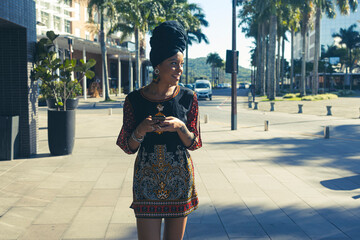 Beautiful African American woman wearing an african turban standing up texting on the mobile cell phone and looking to the side. Palm trees and city buildings on the background.