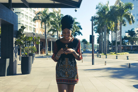Beautiful African American woman wearing casula red and black dress and african turban standing up texting on the mobile cell phone and smiling. Palm trees and city buildings on the background.