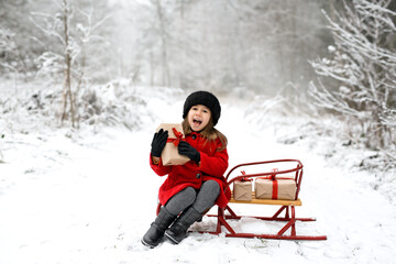 Fototapeta na wymiar A laughing five-year-old girl is sitting on a sled in a winter forest on a cold snowy day with a Christmas gift in her hands. It's time to accept and give gifts. Waiting for the New Year