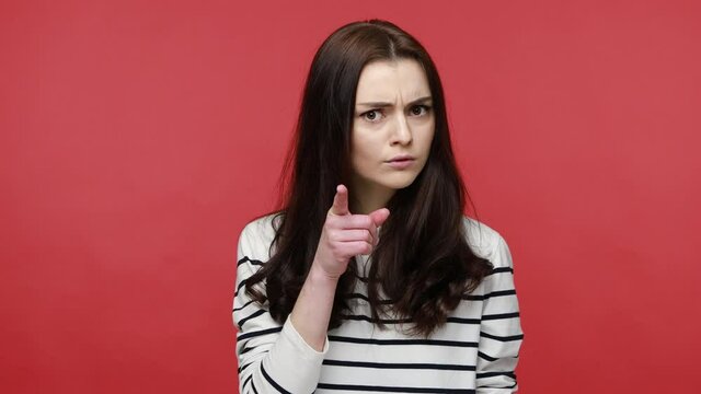 Portrait of strict young adult woman seriously pointing finger and looking at camera, warning, wearing casual style long sleeve shirt. Indoor studio shot isolated on red background.