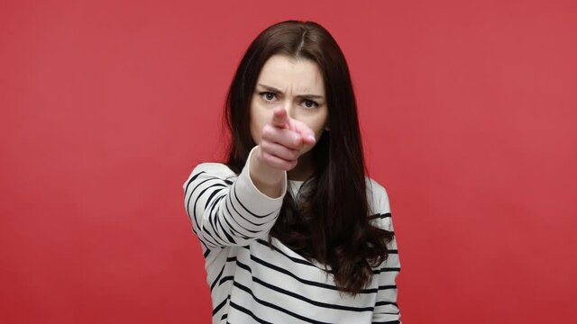 Woman pointing at camera with angry expression, accusing blaming, indicating at you, making choice, wearing casual style long sleeve shirt. Indoor studio shot isolated on red background.