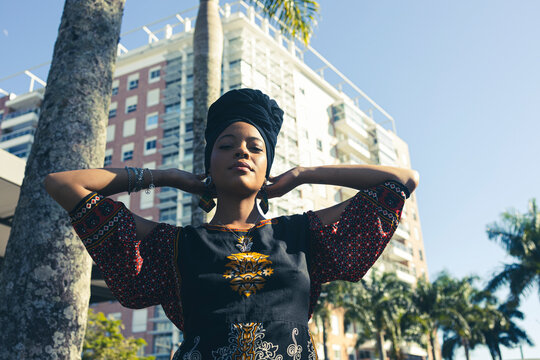 Beautiful African American woman wearing an african turban standing up with her hands behind her head looking down at the camera. Palm trees and city buildings on the background.