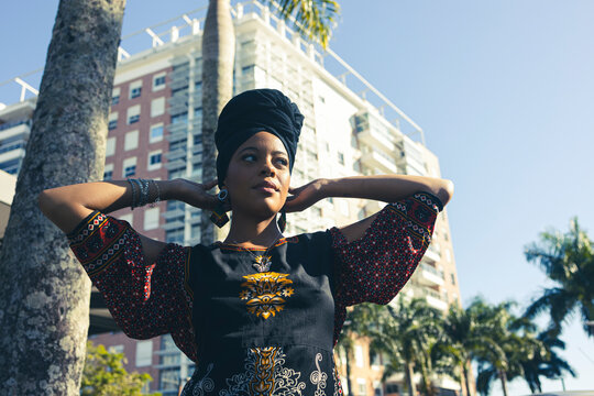 Beautiful African American woman wearing an african turban standing up with her hands behind her head looking away to the side. Palm trees and city buildings on the background.