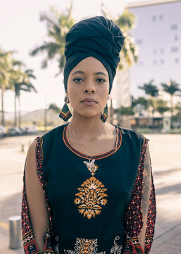 Beautiful African American woman wearing an african turban standing up on the broadwalk looking seriously at the camera. Palm trees and city buildings on the background.