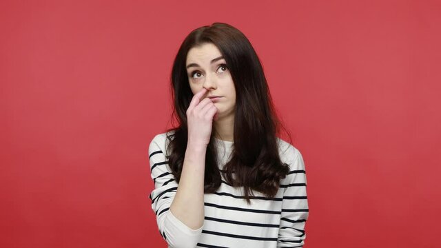 Portrait of brunette woman holding finger in her nose, uncultured bored girl having fun, bad manners, wearing casual style long sleeve shirt. Indoor studio shot isolated on red background.