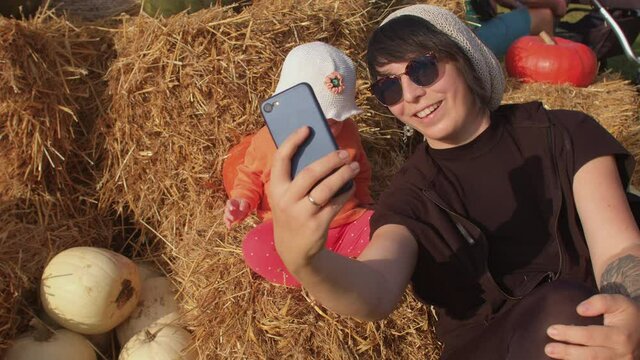 Mom with a child takes a selfie in the countryside, using phone for take a picture. Hay and pumpkins on the background.