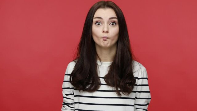 Portrait of woman making fish face grimace with pout lips, looking with confused comical expression, wearing casual style long sleeve shirt. Indoor studio shot isolated on red background.