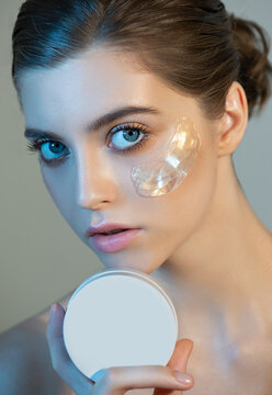 Beautiful Young Woman With Light Makeup And Gold Eye Patches Mask On Her Face Holds The Cream Jar. Portrait Of Beauty Model With Natural Make-up Cares About Her Skin. Spa, Skincare And Wellness