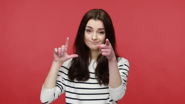 Woman expressing disrespect, showing L finger sign, loser or lame gesture and pointing to camera, wearing casual style long sleeve shirt. Indoor studio shot isolated on red background.