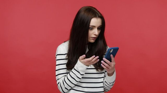 Woman scrolling social media using cell phone, becoming surprised amazed by mobile application, wearing casual style long sleeve shirt. Indoor studio shot isolated on red background.