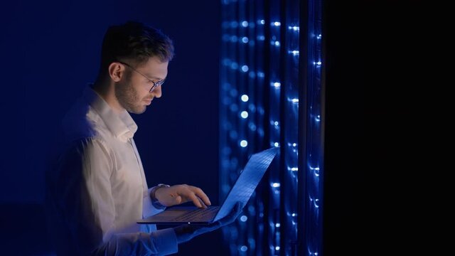 Data center IT specialists coworking in secure server room of cyberspace. Male administrator pointing at error server cabinet running diagnostics on laptop computer