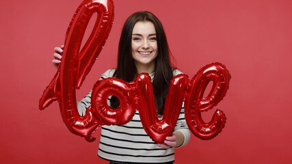Happy woman holding love word from foil ballons, expressing romantic feelings, sending air kissing, wearing casual style long sleeve shirt. Indoor studio shot isolated on red background.
