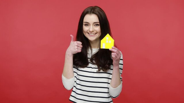 Woman holding paper house and smiling to camera, advertising real estate agency, home purchase, wearing casual style long sleeve shirt. Indoor studio shot isolated on red background.