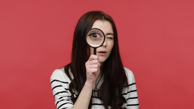 Portrait of woman looks into camera through magnifying glass, inspects something, private detective, wearing casual style long sleeve shirt. Indoor studio shot isolated on red background.