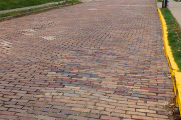 Undulating and settled road surface of old red brick, repairs in odd colored bricks, bright yellow curb, horizontal aspect