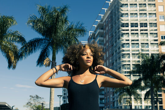 Beautiful African American woman tiding up her afro hair wearing a casual black dress looking away. City buildings and palm trees on the background.