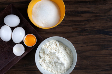 White eggs with one open showing the yolk, small plate with sugar and one with flour, on a dark background. Space for text.