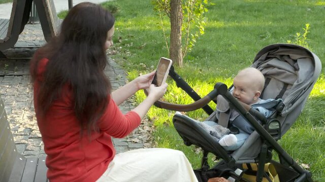 Mother photographing baby in stroller with smartphone outdoors, cute caucasian six month old child. High quality 4k footage