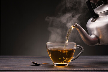 Pour hot tea into a teacup placed on a wooden table with, dark black background
