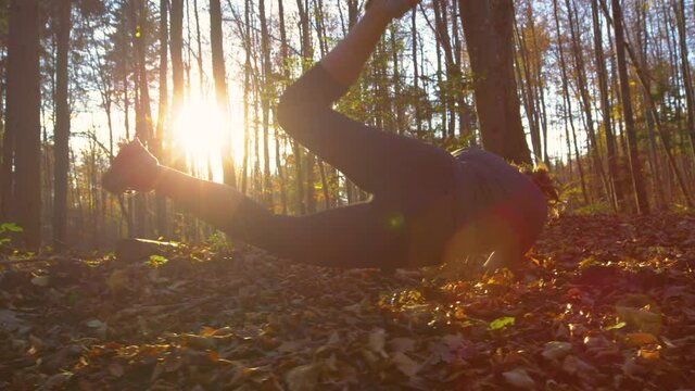 SLOW MOTION, LENS FLARE, LOW ANGLE: Young Caucasian woman running through the fall colored woods at sunset trips and falls into heap of leaves. Female jogger stumbles to ground while running in forest
