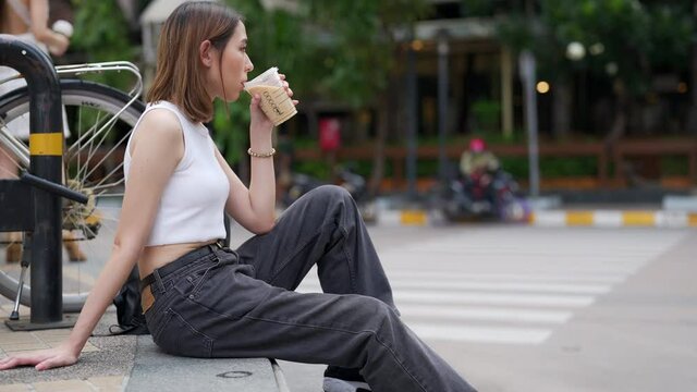 Portrait of Young beautiful Asian woman in casual clothing sitting on outdoor bench in the city and drinking iced coffee with looking crowd of people. Pretty girl enjoy urban lifestyle and city life.