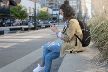 Obraz premium Woman seated with mask and mobile phone texting with backpack hanging down
