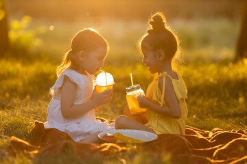 Two little mixed race girls drink smoothies in the garden outdoors, sitting on a blanket on a warm sunny summer day. Healthy snack for children. GMO free food. Concept of a healthy lifestyle. 