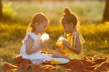 Two little mixed race girls drink smoothies in the garden outdoors, sitting on a blanket on a warm sunny summer day. Healthy snack for children. GMO free food. Concept of a healthy lifestyle. 