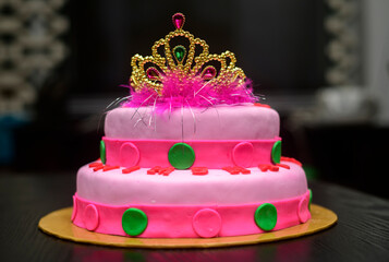 Two-tier fondant cake design with a tiara on top, decorated with icing flowers. Delicious pink-red color-themed round cake on yellow cake tray against the dark background.