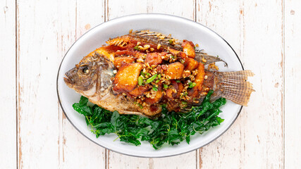 tasty large fried nile tilapia fish with chili, garlic and crispy holy basil leaves in oval ceramic plate on white wooden texture background, top view