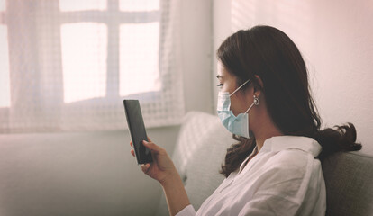 Close-up of an Asian woman wearing a face mask and online searching on a cellphone at home on a couch. New Business home office during Coronavirus or Covid-19 quarantine concept