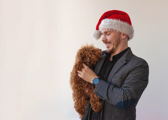 A young man in a Santa Claus hat holds a miniature poodle dog in his arms